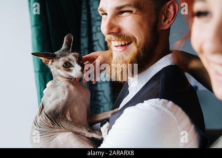 Newly wed couple standing next to the window, cuddling their hairless sphynx cat Stock Photo