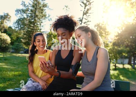 Diverse sporty young female friends sitting together on bench looking at mobile phone in the park at morning - happy fitness friends smiling and Stock Photo