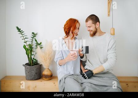Happy husband and wife sitting together on a shelf, drinking coffee at home. Stock Photo