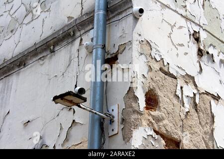 Three new closed circuit TV CCTV security cameras mounted on corner of old abandoned building with dilapidated cracked facade next to new gutter pipe Stock Photo