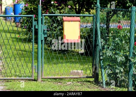 Yellow and red mailbox with faded color mounted on old green metal and wire mesh fence surrounding suburban family house garden filled with flowers Stock Photo