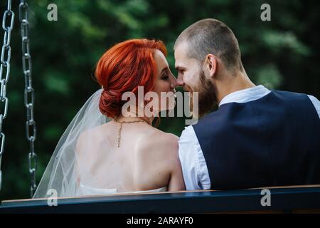 Happy newly wed couple sitting on swing, touching noses. from behind Stock Photo