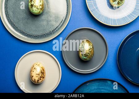 Different Ceramic Plates with Golden Speckled Easter Eggs of pastel colors on blue background. Happy Easter card concept, minimalistic design, top view Stock Photo
