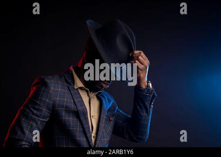 Side view of cool dark skinned man in stylish suit, touching his black hat, looking down. Closeup portrait of fashionable african american male model Stock Photo