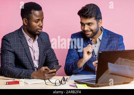Two young diverse business people chatting sitting in office, using laptop and cellphone. Indian man sharing ideas or business plan with dark skinned Stock Photo