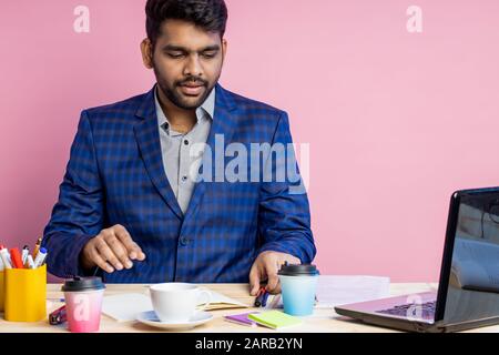 Stylish serious handsome young indian businessman sitting at desk in office, wearing checked suit, holding smartphone, checking email, using mobile ba Stock Photo