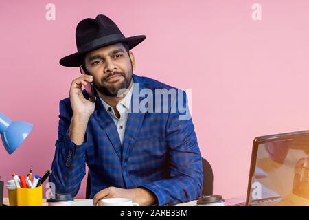 Joyful stylish indian entrepreneur wearing formal wear, black hat, talking on mobile phone, smiling broadly, looking to side, sitting at desk in offic Stock Photo