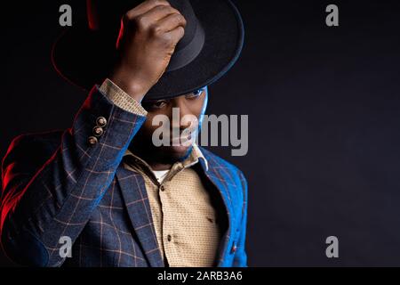 Cool dark skinned man in stylish suit, touching his black hat, looking down. Closeup portrait of fashionable african american male model posing on dar Stock Photo
