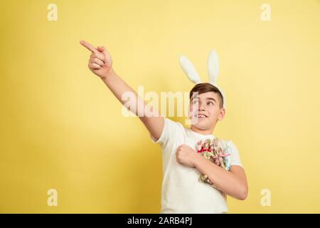 Astonished pointing up. Decorating. Caucasian boy as an Easter bunny on yellow background. Happy easter greetings. Beautiful male model. Concept of human emotions, facial expression, holidays. Copyspace. Stock Photo