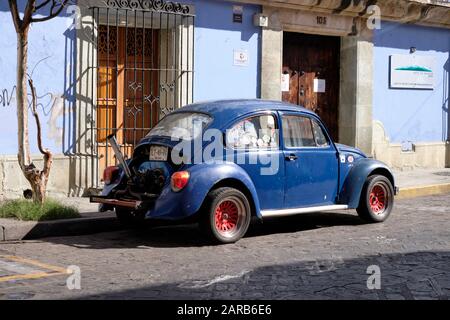 Blue classic Volkswagen beetle car parked on street next to a colonial house, painted also blue.  The engine has been modified with the hood removed Stock Photo