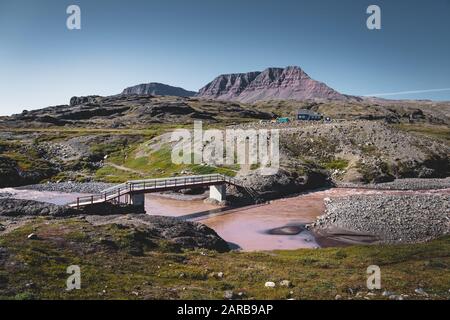 Hiking path on arctic Disko island in Greenland. River bed with bridge and colourful houses in background. Table mountains on a sunny day in summer. Stock Photo