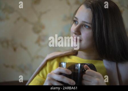 A picture of nice and thoughtful girl sits at sofa and drinks tea. She has brown blanket on her shoulders. Woman got cold. She suts at window Stock Photo