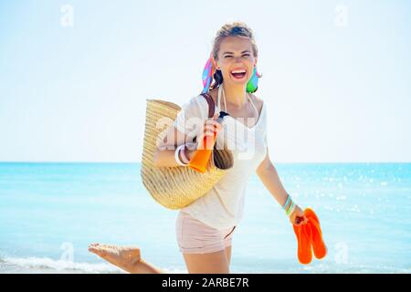 Portrait of cheerful middle age woman in white t-shirt with beach straw bag, orange flip flops and bottle of sunscreen on the beach. Stock Photo