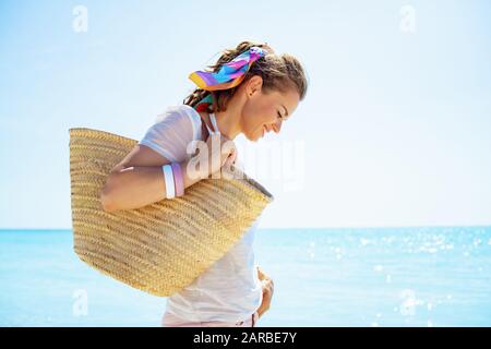 happy modern 40 year old woman in white t-shirt with beach straw bag on the beach. Stock Photo