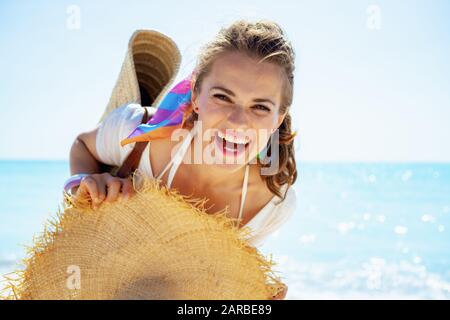 Portrait of happy modern middle age woman in white t-shirt with beach straw bag and big hat on the seashore.
