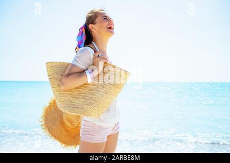 happy trendy woman in white t-shirt with beach straw bag and big hat on the seashore. Stock Photo