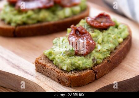 Vegetarian food. Rye bread with guakomole, avocado pasta and dried tomatoes, on wooden cutting board. Avocado toast. Stock Photo
