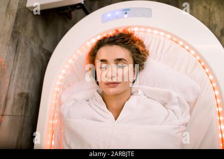 Top view of the face of pretty woman with closed eyes, relaxing while lying in modern hot spa treatment capsule in cosmetology clinic Stock Photo