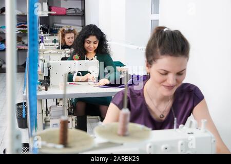 Young dressmaker woman sews clothes on sewing machine. Smiling seamstress and her hand close up in workshop. Focus on sewing machine and tissue. Stock Photo