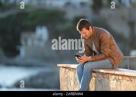 Adult man using mobile phone sitting on a wall of a balcony in a coast town Stock Photo