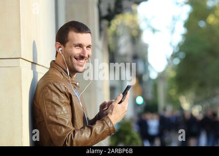 Happy adult man wearing earphones holding mobile phone looks at you in the street Stock Photo