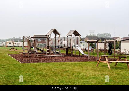 The climbing frames and slides of a children's play area stand empty on a woodchip safety surface under a grey sky as sea mist envelops the distance Stock Photo