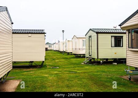 Rows of light coloured static caravans stand on concrete pads surrounded by neatly mown green grass under a greyish sky as sea mist obscures the sun Stock Photo