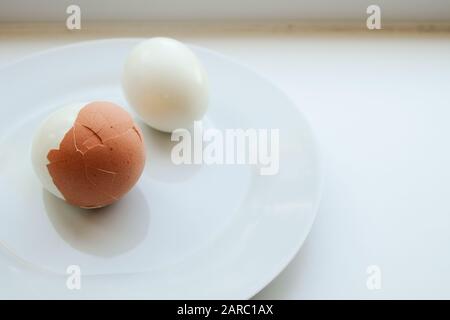 Two boiled eggs on a white plate on a white background. One egg peeled the other with its shell partly removed. Stock Photo