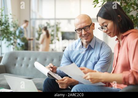 Portrait of bald mature businessman smiling at Asian young woman while discussing project in modern office, copy space Stock Photo