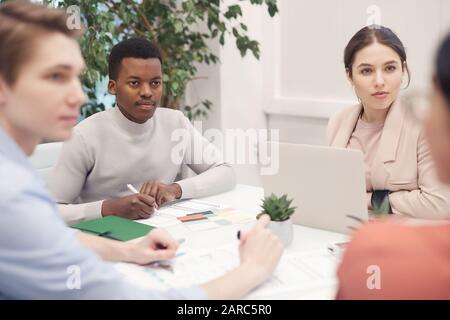 Multi-ethnic group of business people discussing project while working sitting at meeting table, focus on African-American man listening intently to p Stock Photo