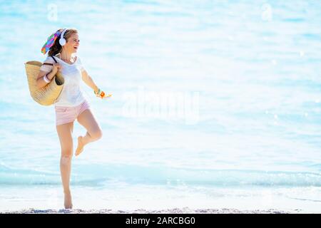 happy trendy middle age woman in white t-shirt and pink shorts with beach straw bag holding orange flip flops, listening to the music with headphone a Stock Photo