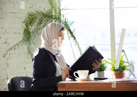 Reviewing of documents. Portrait of a beautiful arabian businesswoman wearing hijab while working at openspace or office. Concept of occupation, freedom in business area, success, modern solution. Stock Photo