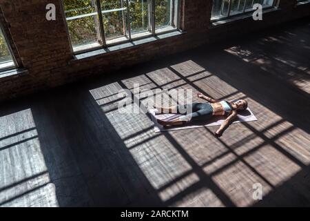 Young calm woman practicing yoga lying in dead body pose relax on mat in studio