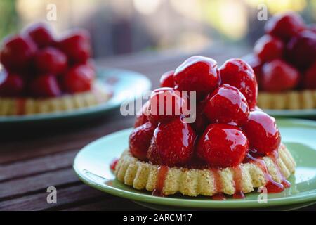 Three fresh vegan strawberry tarts, served on green plates to a wooden table on the balcony - side view, horizontal orientation, blurred background Stock Photo