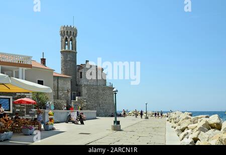 Piran, Slovenia - July 4th 2015: Unidentified people on promenade and medieval church Sveti Klementa in the picturesque village on Adriatic sea Stock Photo