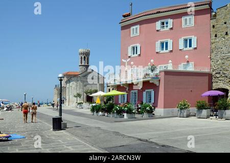 Piran, Slovenia - July 4th 2015: Unidentified people on beach and promenade in the picturesque village on Adriatic sea, medieval church Saint Clementa Stock Photo