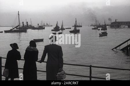 1940s, historical, father, mother and daughter in Gravesend, Kent, England, UK, looking at the harbour and the moored boats including a steamship. Until the building of the Tilbury docks in 1886, Gravesend was the Thames's first port of entry. The Royal Terrace Pier can be seen in the top right of the picture. Stock Photo