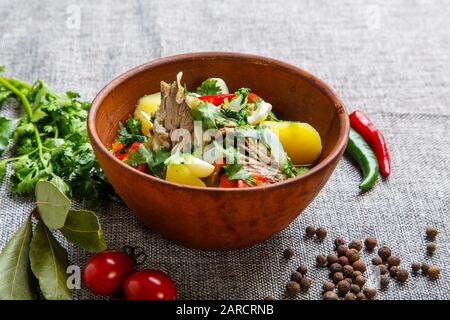 Hashlama in a clay plate. Ingredients: Beef ribs, potatoes, herbs, garlic. The plate stands on a textured gray canvas. Around lie pepper red and green Stock Photo