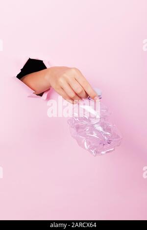 Female hand holds through a hole a empty crumpled plastic bottle on a pink background, no plastic and zero waste concept Stock Photo