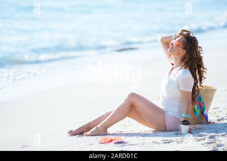 relaxed young woman in white t-shirt and pink shorts with coffee cup sitting on the ocean shore. Stock Photo