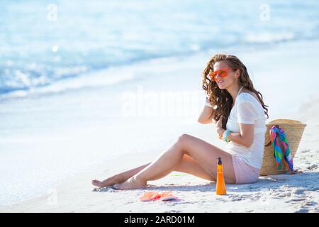 modern woman in white t-shirt and pink shorts on the ocean coast adjusting long wavy hair while sitting. Stock Photo