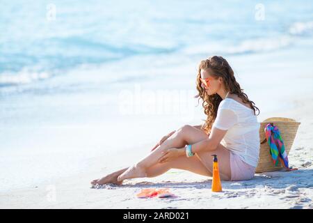 trendy middle age woman in white t-shirt and pink shorts checking legs while sitting on the seashore. Stock Photo