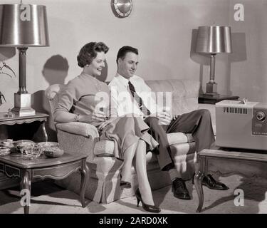 1950s MAN AND WOMAN SMILING SITTING ON COUCH WATCHING A SMALL PORTABLE TELEVISION DRINKING GLASSES OF BEER - f5571 HAR001 HARS COUCH STYLE RELAXING LIFESTYLE FEMALES MARRIED SPOUSE HUSBANDS HOME LIFE COPY SPACE FRIENDSHIP HALF-LENGTH LADIES PERSONS MALES ENTERTAINMENT PORTABLE B&W PARTNER DATING TELEVISIONS HAPPINESS BROADCASTING LEISURE AND WHITE SHIRT ATTRACTION NECKTIE CONNECTION COURTSHIP STYLISH END TABLE POSSIBILITY CHANNELS COOPERATION MID-ADULT MID-ADULT MAN MID-ADULT WOMAN REFRESHING SOCIAL ACTIVITY TABLE LAMPS TOGETHERNESS WIVES BLACK AND WHITE CAUCASIAN ETHNICITY COFFEE TABLE Stock Photo