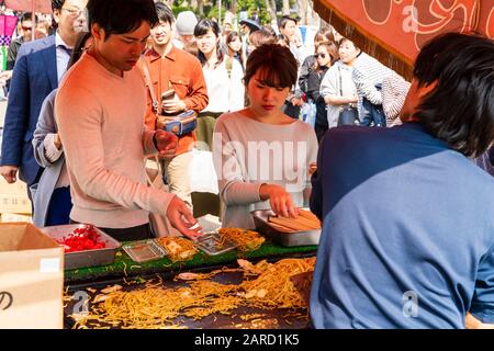 Over the shoulder view of stall holder selling plastic box of hot noodles to young couple with many others queuing behind at Osaka Castle Park, Japan. Stock Photo