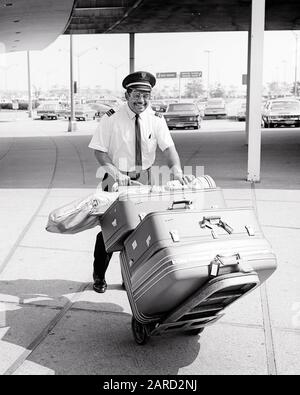 1960s SMILING AFRICAN-AMERICAN MAN AIRPORT PORTER PUSHING CART FULL OF LUGGAGE AT CURBSIDE - s17675 HAR001 HARS FULL-LENGTH PERSONS INSPIRATION MALES CONFIDENCE TRANSPORTATION MIDDLE-AGED B&W MIDDLE-AGED MAN EYE CONTACT SKILL OCCUPATION HAPPINESS SKILLS CHEERFUL CUSTOMER SERVICE AFRICAN-AMERICANS AFRICAN-AMERICAN BLACK ETHNICITY LABOR PRIDE OPPORTUNITY EMPLOYMENT OCCUPATIONS SMILES CONNECTION JOYFUL SUPPORT CURBSIDE EMPLOYEE GOLF CLUBS COOPERATION PORTER BAGGAGE BLACK AND WHITE HAR001 LABORING OLD FASHIONED AFRICAN AMERICANS Stock Photo