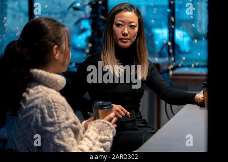 Caucasian And Asian Young Women Talking In Cafe And Drinking Coffee. Concept Of Multiethnic And International Friendship. Stock Photo