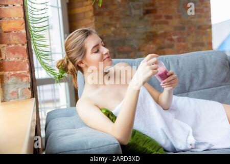 Beauty Day for yourself. Long haired woman wearing towel doing her daily skincare routine at home. Sits on sofa, delighted using rose pink sea's salt. Concept of beauty, self-care, cosmetics. Stock Photo