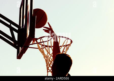 Young silhouetted basketball player jumping high to the target basketball hoop achieves to score with a flying slam dunk at sunset. Success and target Stock Photo