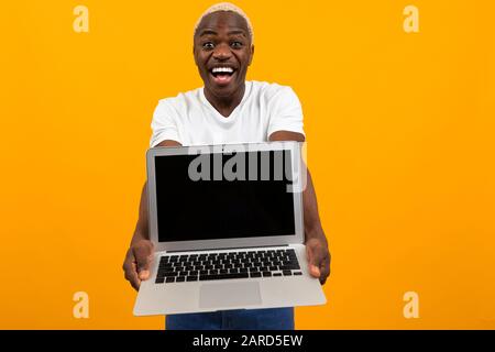 attractive joyful surprised american man holds out his hands with laptop with mockup on yellow background. Stock Photo