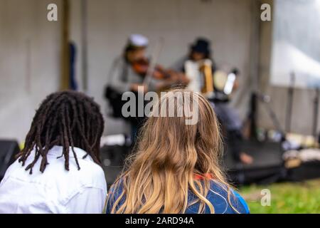 Closeup rear view of woman with dreadlocks hair by female friend looking at artist performing guitar and accordion in event at outdoor Stock Photo